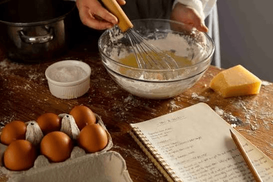 Tangan mengocok adonan di mangkuk kaca, dengan telur, keju, dan buku catatan resep di meja dapur.
